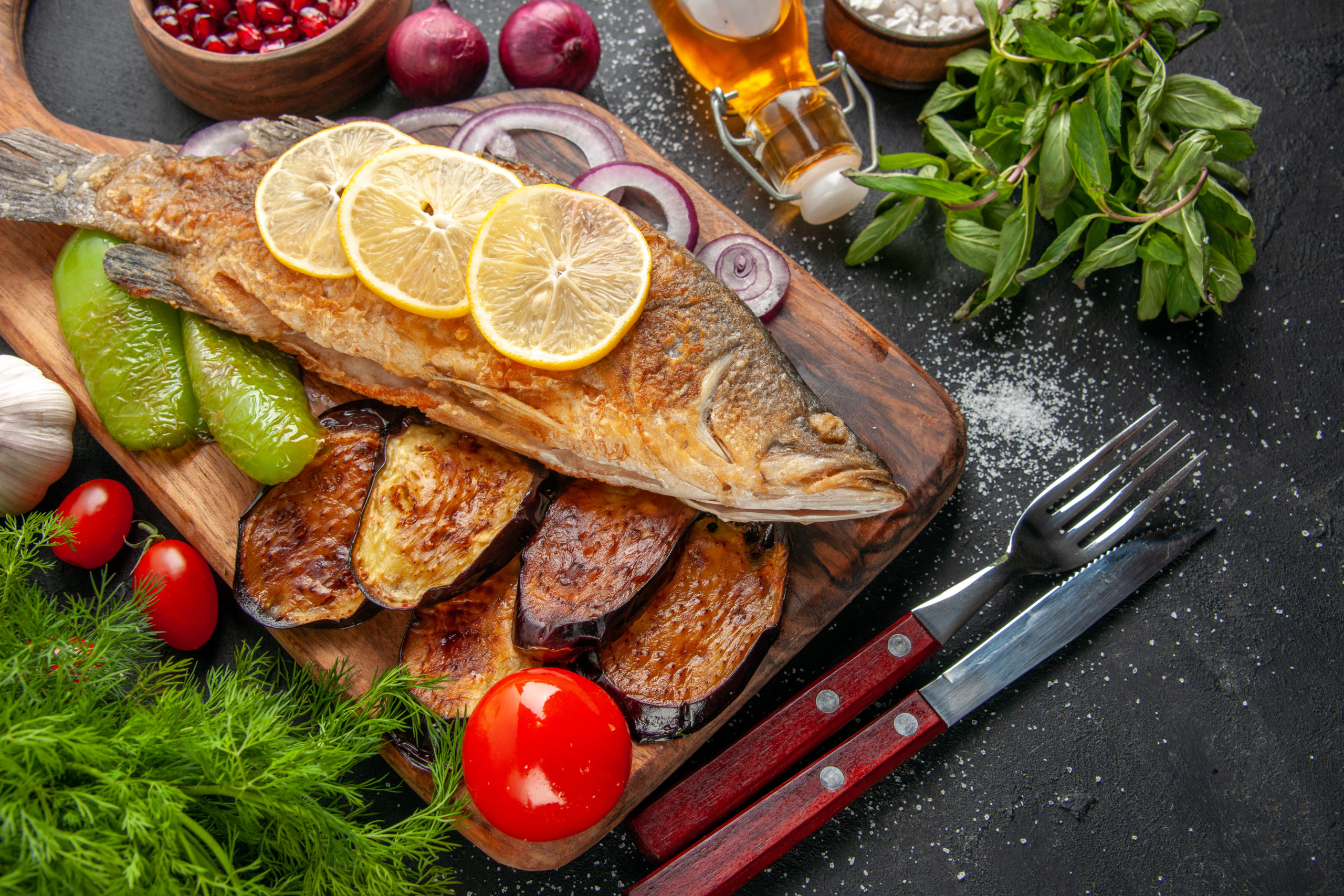 bottom-view-fish-fry-fried-eggplants-onion-peppers-on-wood-board-spices-in-small-bowls-fork-and-knife-tomatoes-oil-bottle-mint-dill-on-dark-background